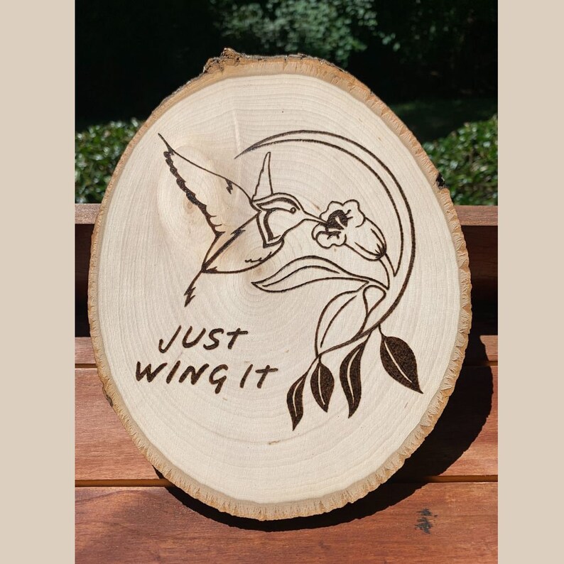 May include: A round wooden plaque with a hummingbird and floral design, pyrographed onto the wood. The text "JUST WING IT" is below the hummingbird. The wood has a natural, light color and a visible wood grain.