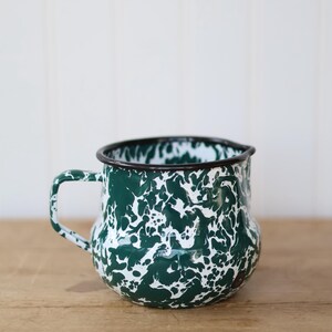 May include: A green and white enamel mug with a speckled pattern. The mug has a black rim and handle, and sits on a wooden surface. This rustic mug is perfect for coffee, tea, or other beverages.
