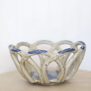 May include: A ceramic basket with a cream-colored base and blue accents. The basket has an open, woven design with a scalloped rim. The speckled texture adds a rustic touch to this decorative piece.