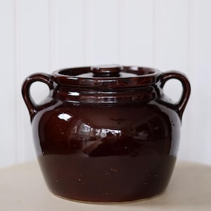 May include: A dark brown ceramic crock pot with a lid and two side handles. The pot has a glossy finish and is set against a white background. This kitchenware item is suitable for cooking and food storage.