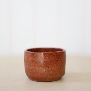 May include: A small, round, handmade ceramic bowl with a textured, reddish-brown exterior. The bowl has a simple, minimalist design, perfect for serving dips or small portions. The interior is a darker shade of brown.