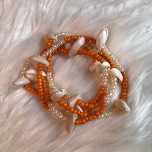 Op de afbeelding: Een gelaagde armband gemaakt met oranje en witte kralen en witte schelpen.