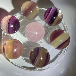 May include: A selection of polished gemstone spheres in purple, yellow, and pink hues. The spheres are displayed on a clear, faceted glass stand. One pink sphere features a star pattern. These gemstones are likely used for decorative or spiritual purposes.