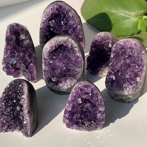 Op de afbeelding: Een verzameling amethistkristalgeodes in verschillende maten en vormen. De geodes hebben een dieppaarse kleur met kristallijne interieurs en een ruwe, grijze buitenkant. De opstelling omvat verschillende rechtopstaande en afgeronde formaties, die de natuurlijke schoonheid laten zien.