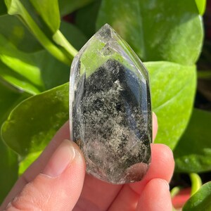 Pode incluir: Um cristal de quartzo transparente com inclusões verde escuro. O cristal é segurado em uma mão, com folhagem verde ao fundo. O cristal tem uma forma alongada polida com uma ponta.