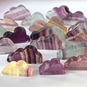 May include: A collection of cloud-shaped decorative objects in various colors, including purple, pink, yellow, and green. The objects appear to be made of a translucent, polished material, possibly a type of stone or crystal. The clouds have a smooth, rounded shape.