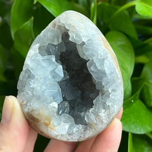 May include: A light blue crystal geode with a hollow center, revealing a dark gray interior. The geode is smooth and rounded, with a natural, earthy texture.