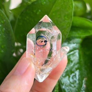Pode incluir: Uma ponta de cristal de quartzo transparente de terminação dupla com fraturas internas e uma pequena marca circular azul distinta na superfície. O espécime mineral translúcido exibe inclusões naturais e facetas em toda a estrutura.