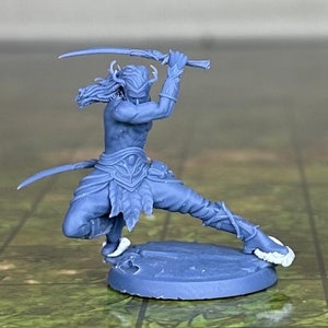 Op de afbeelding: Een blauwe plastic miniatuurfiguur van een elfenkrijger die twee zwaarden vasthoudt, staand op een ronde basis. De elf draagt ​​een harnas en heeft lang haar. Een gouden en zwarte 20-zijdige dobbelsteen bevindt zich op de achtergrond.