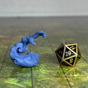 Puede incluir: Una miniatura de plástico azul de una mano etérea que gira y se estira hacia arriba. La mano está colocada sobre una superficie con un patrón verde y marrón con un dado de 20 caras negro y dorado en el fondo.