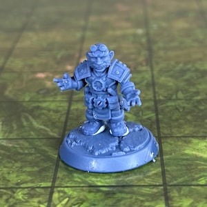 May include: A blue plastic miniature figurine of a dwarf wearing goggles, armor, and a belt with a gear. The dwarf is holding a tool in one hand and pointing with the other. The figurine is standing on a round base.