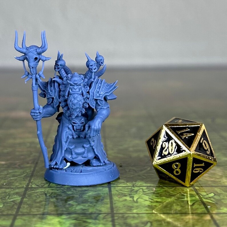 Puede incluir: Una miniatura de pl&aacute;stico azul de un personaje de fantas&iacute;a que sostiene un bast&oacute;n con calaveras. El personaje lleva armadura y tiene barba. La miniatura est&aacute; de pie sobre una base redonda. Un dado de 20 caras negro y dorado est&aacute; en el fondo.