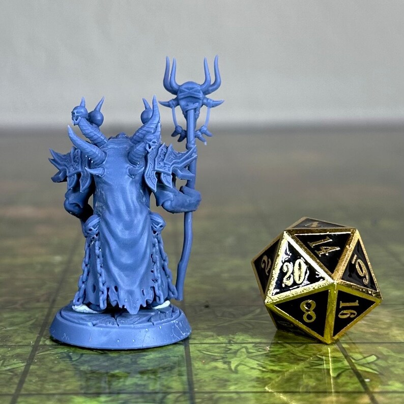 Puede incluir: Una figura en miniatura de pl&aacute;stico azul de un demonio sosteniendo un bast&oacute;n con una calavera en la parte superior. La figura est&aacute; de pie sobre una base redonda. Un dado de 20 caras de oro est&aacute; en el fondo.