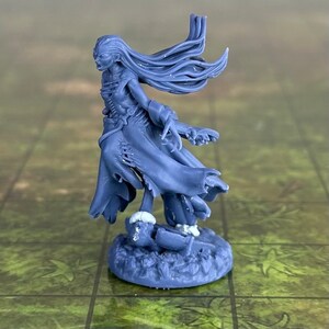 Puede incluir: Una figura en miniatura de plástico azul de una figura femenina con el pelo largo y suelto, que lleva una túnica desgarrada y está de pie sobre una base con una calavera rota.