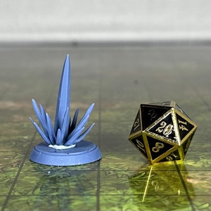 Puede incluir: Una formación de cristal azul claro impresa en 3D y un dado de veinte caras negro y dorado. El cristal tiene una base circular y una espiga central puntiaguda con púas más pequeñas. El dado tiene números y detalles dorados.