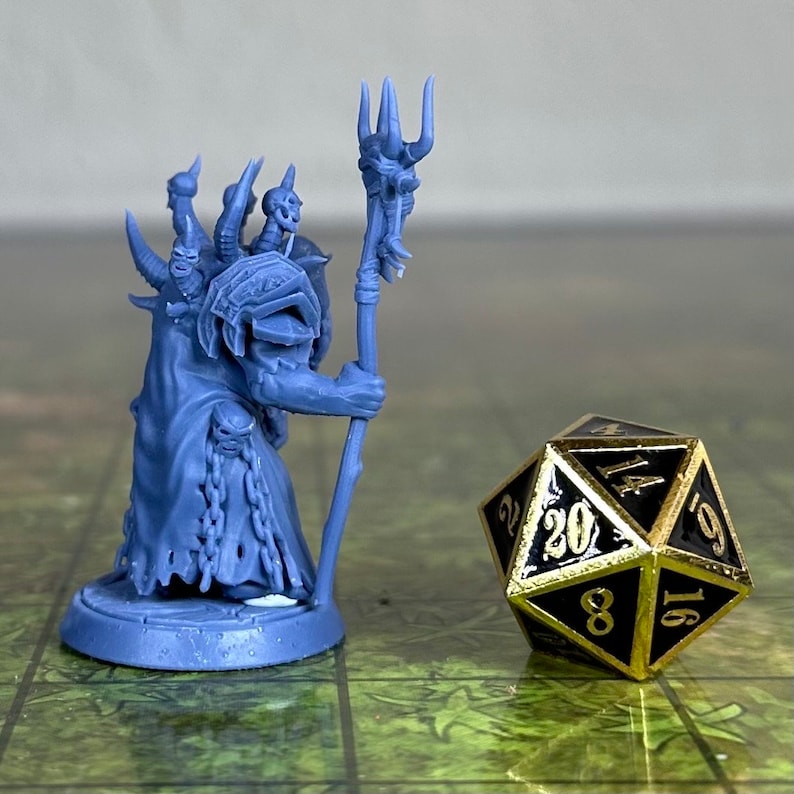 Puede incluir: Una figura en miniatura de pl&aacute;stico azul de una criatura fant&aacute;stica que sostiene un bast&oacute;n con una calavera en la parte superior. La figura est&aacute; de pie sobre una base redonda. Un dado de 20 caras negro y dorado est&aacute; en primer plano.