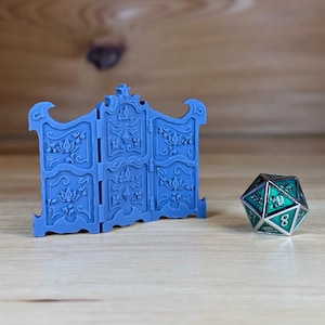 May include: A blue, three-panel folding screen with intricate floral designs. The screen is made of plastic and is about 6 inches tall. A green and silver metal dice is in the foreground.