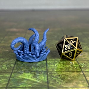 May include: A blue miniature octopus emerging from water next to a black and gold twenty-sided die. The die has white numbers and a gold trim. The octopus is a detailed figurine, likely for tabletop gaming.