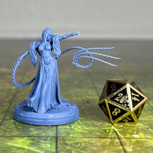 May include: A light blue miniature figure of a woman in a flowing gown, holding a whip, next to a black and gold twenty-sided die. The figure is on a round base, and the die has gold trim and black numbers.