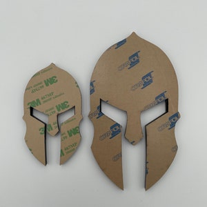 Op de afbeelding: Twee Spartanen helm sjablonen, een groter dan de andere, zijn gesneden uit bruin papier. De sjablonen zijn bedekt met een transparante kleefrug. De kleefrug heeft de tekst "3M 467MP 200MP Adhesive" erop gedrukt. De sjablonen zijn ook bedekt met een bruine papieren rug met de tekst "ORAFOL" erop gedrukt.