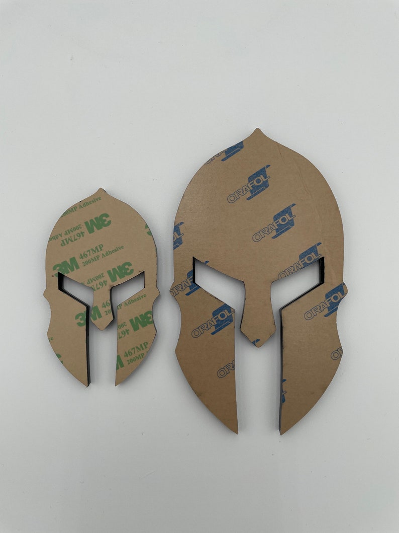 Op de afbeelding: Twee Spartanen helm sjablonen, een groter dan de andere, zijn gesneden uit bruin papier. De sjablonen zijn bedekt met een transparante kleefrug. De kleefrug heeft de tekst "3M 467MP 200MP Adhesive" erop gedrukt. De sjablonen zijn ook bedekt met een bruine papieren rug met de tekst "ORAFOL" erop gedrukt.
