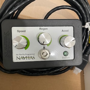 May include: A black control box with three knobs labeled "Speed", "Regen", and "Accel". The box also has a green status light and a keyhole. The text "On The Fly Programmer NAVITAS" is printed on the box.