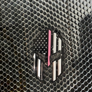Op de afbeelding: Een zwart-witte Spartaanse helm met een dunne roze streep in het midden, gesuperponeerd op een Amerikaans vlagontwerp. De helm bevindt zich op een zilveren metalen gaasachtergrond.
