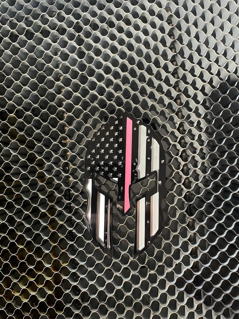 Op de afbeelding: Een zwart-witte Spartaanse helm met een dunne roze streep in het midden, gesuperponeerd op een Amerikaans vlagontwerp. De helm bevindt zich op een zilveren metalen gaasachtergrond.