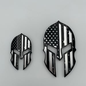 Op de afbeelding: Twee zwarte en zilveren Spartan helm stickers met een Amerikaans vlag ontwerp. De grotere sticker is ongeveer 10 cm hoog en de kleinere ongeveer 5 cm hoog.