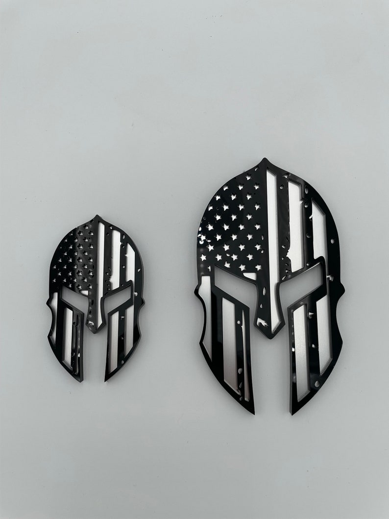 Op de afbeelding: Twee zwarte en zilveren Spartan helm stickers met een Amerikaans vlag ontwerp. De grotere sticker is ongeveer 10 cm hoog en de kleinere ongeveer 5 cm hoog.