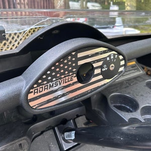 May include: A black steering wheel cover with a silver American flag design and the text "ADAMSVILLE" printed on it. The cover is installed on a golf cart.