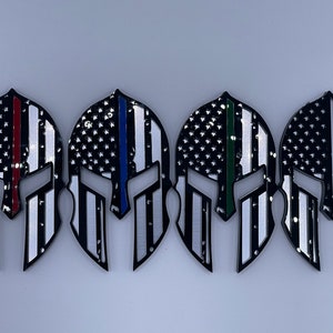 Op de afbeelding: Vier zwarte Spartanen helm stickers met een Amerikaans vlag ontwerp. Elke helm heeft een dunne gekleurde streep die een andere groep eerstehulpverleners vertegenwoordigt: rood voor brandweerlieden, blauw voor politie, groen voor ambulancepersoneel en zwart voor militairen.