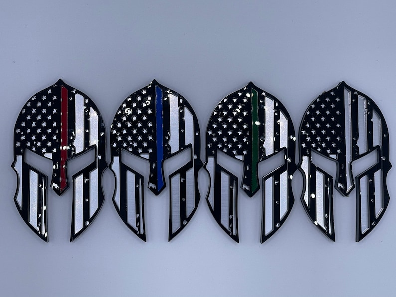 Op de afbeelding: Vier zwarte Spartanen helm stickers met een Amerikaans vlag ontwerp. Elke helm heeft een dunne gekleurde streep die een andere groep eerstehulpverleners vertegenwoordigt: rood voor brandweerlieden, blauw voor politie, groen voor ambulancepersoneel en zwart voor militairen.