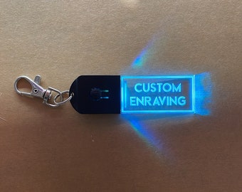 May include: A black keychain with a clear acrylic insert that lights up blue. The acrylic insert has the text "CUSTOM ENGRAVING" engraved on it.