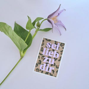 以下が含まれることがあります： 緑の葉の小枝と、ヒョウ柄の背景にラベンダー色の文字で「sei lieb zu dir」と書かれたカードの隣にある薄紫色の花。画像は白い背景に設定されています。
