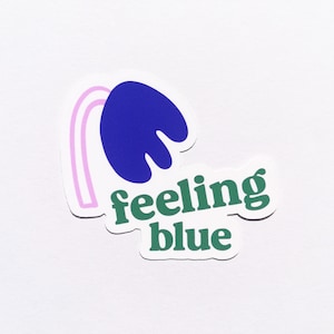 Könnte beinhalten: Ein Aufkleber mit einer blauen abstrakten Form und einer rosa gebogenen Linie. Die Worte "feeling blue" sind grün, mit weißer Umrandung. Der Aufkleber befindet sich auf weißem Hintergrund.