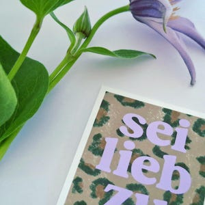 以下が含まれることがあります： ヒョウ柄の背景に、ラベンダー色の文字で「Sei lieb zu dir」と書かれたグリーティングカード。緑の葉の小枝と紫の花が背景にあります。
