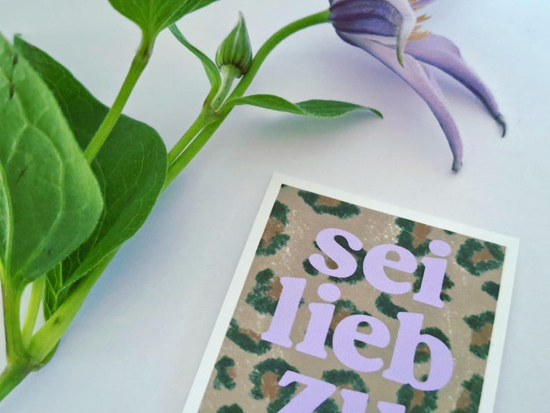 以下が含まれることがあります： ヒョウ柄の背景に、ラベンダー色の文字で「Sei lieb zu dir」と書かれたグリーティングカード。緑の葉の小枝と紫の花が背景にあります。