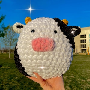 May include: A handmade, round crochet cow plushie. The plushie is white with black patches, a pink muzzle, and small black eyes. The cow has small yellow horns and black ears. The plushie is held in front of a bright blue sky.