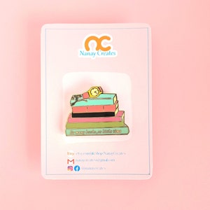 Pode incluir: Um broche de esmalte dourado com uma pilha de livros coloridos com um relógio no topo. Os livros têm o texto "So many books, so little time".