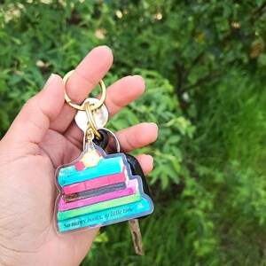 Könnte beinhalten: Ein transparenter Schlüsselanhänger mit einem Stapel bunter Bücher und dem Text "So many books, so little time".