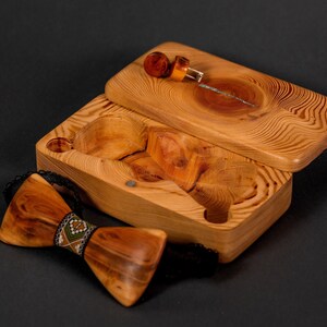 May include: A wooden box with a hinged lid, open to reveal a small wooden bow tie with a colorful patterned band. The box is made of light brown wood with a natural grain pattern.