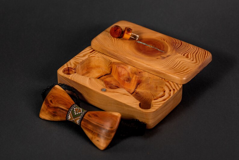 May include: A wooden box with a hinged lid, open to reveal a small wooden bow tie with a colorful patterned band. The box is made of light brown wood with a natural grain pattern.