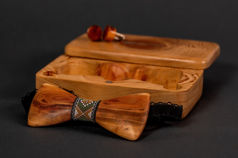 May include: A wooden bow tie with a black and green patterned band in a wooden box with a lid. The box has two small wooden circles inside.