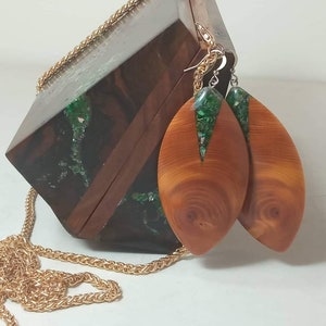 Pode incluir: Uma clutch de caixa de madeira com uma alça de corrente dourada e incrustações de resina verde e marrom. A clutch vem com um par de brincos de madeira com detalhes em resina verde.