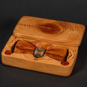 May include: A wooden bow tie in a wooden box. The bow tie is made of light brown wood with dark brown streaks. The box is made of light brown wood with a natural wood grain pattern. The bow tie has a black and green patterned band.