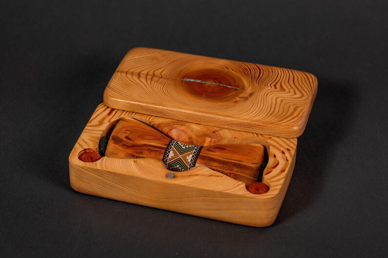 May include: A wooden bow tie in a wooden box. The bow tie is made of light brown wood with dark brown streaks. The box is made of light brown wood with a natural wood grain pattern. The bow tie has a black and green patterned band.