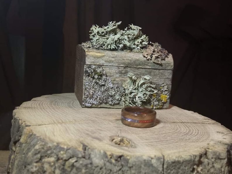 May include: A wooden ring with a blue and red inlay sits on a tree stump next to a small wooden box covered in moss.