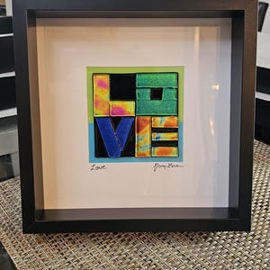 May include: Framed artwork with the word "LOVE" in colorful, textured glass. The letters are arranged in a square, with a black frame. The artwork includes the artist's signature and the word "Love" in cursive.