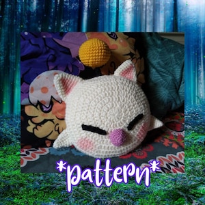 May include: A white crocheted plush toy shaped like a cat with a yellow crocheted ball on top. The toy has pink cheeks and a pink nose. The text "*Pattern*" is written in purple letters on a green background.